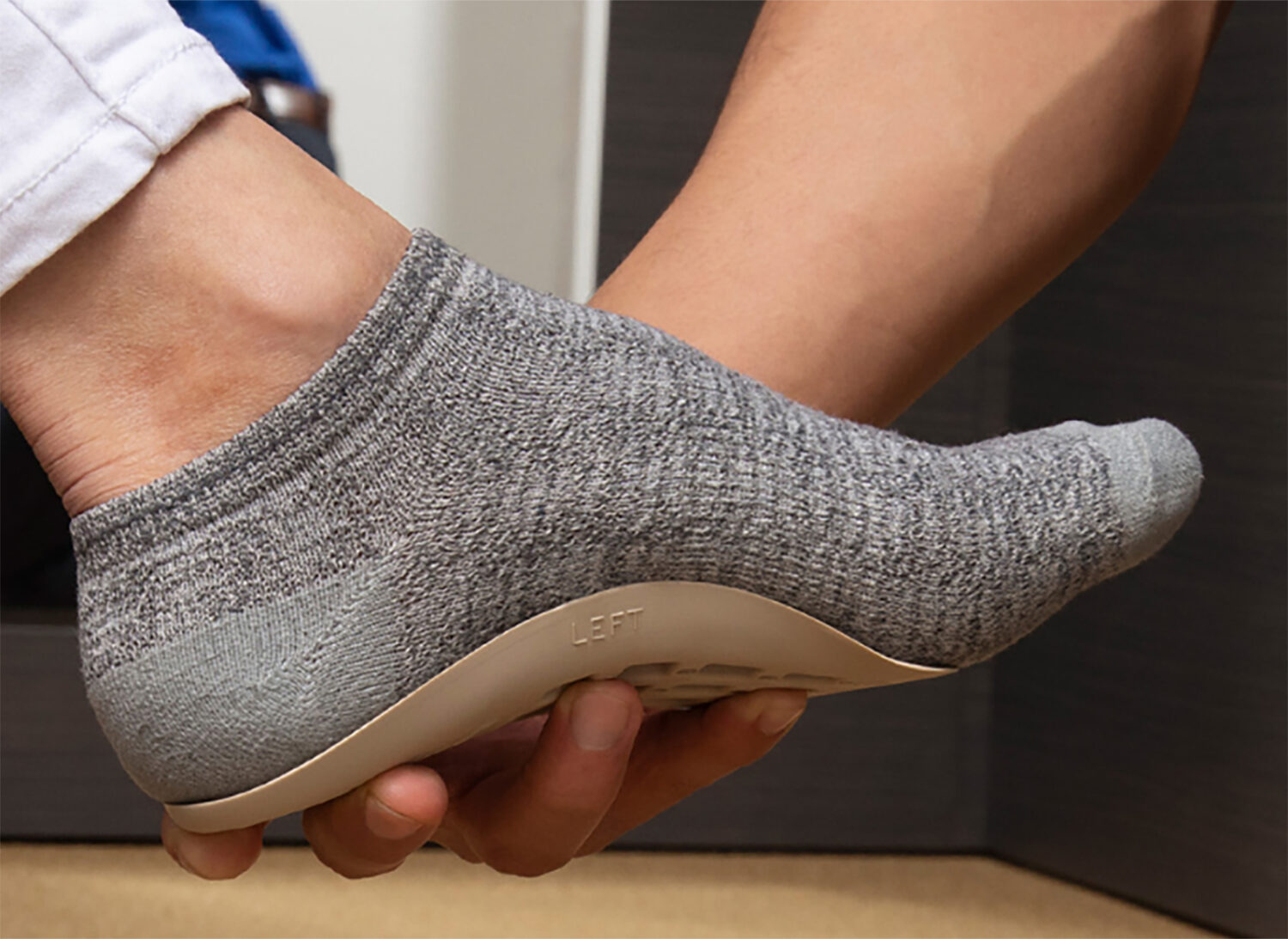A single foot in a grey sock is held by a hand. The foot is supported by a Good Feet Arch Support in between the sock and hand.